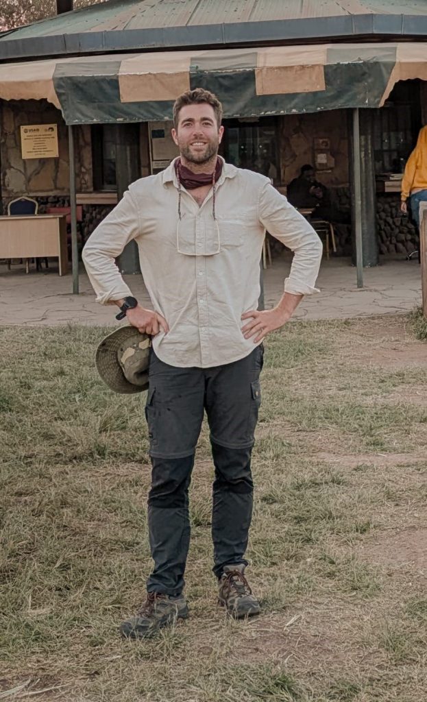 Un hombre de pie al aire libre sobre la hierba, con un sombrero en la mano izquierda, vestido con una camisa beige, pantalones oscuros, botas y una polaina para el cuello, con un edificio al fondo.