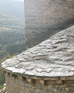 Primer plano de un tejado de tejas de piedra con un borde curvado, con un telón de fondo de colinas y vegetación.