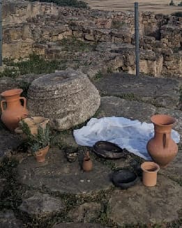 Cerámicas y vasijas de barro están dispuestas sobre suelo de piedra cerca de antiguas ruinas, con un paño blanco parcialmente extendido junto a ellas.
