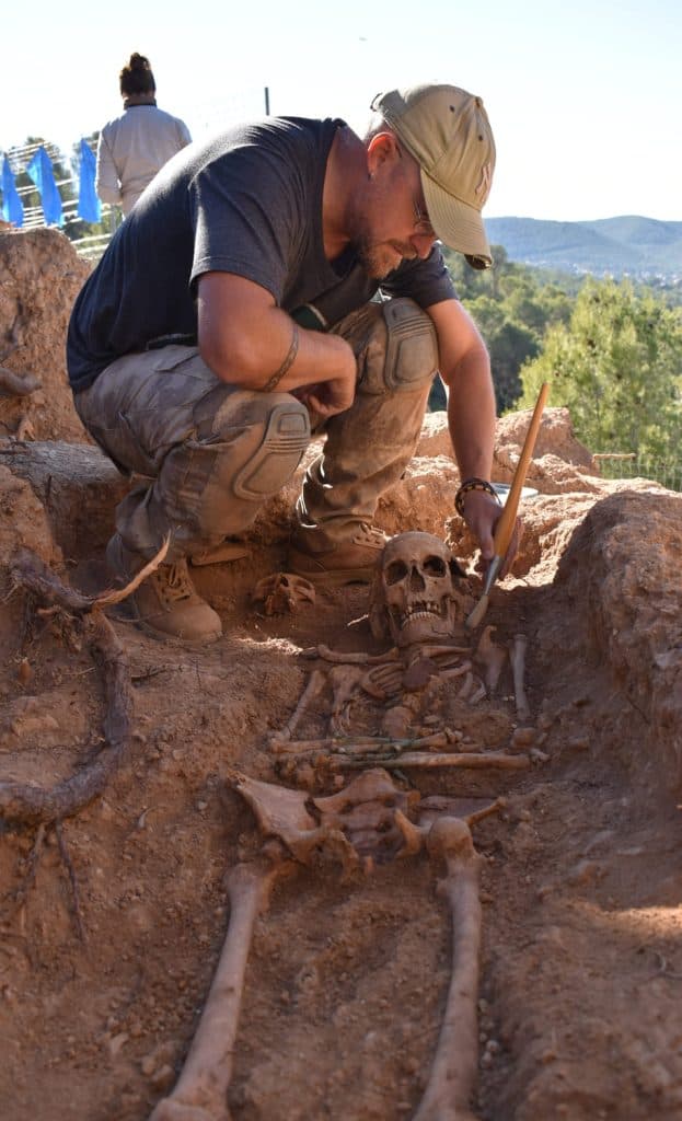 Un arqueólogo se arrodilla junto a un esqueleto humano parcialmente excavado en una excavación al aire libre, cepillando cuidadosamente la suciedad de los huesos.