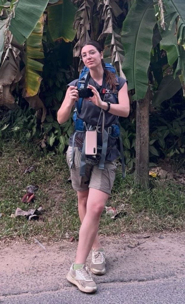 Una persona con equipo de senderismo y una cámara fotográfica junto a una carretera con grandes hojas de plátano verde al fondo.
