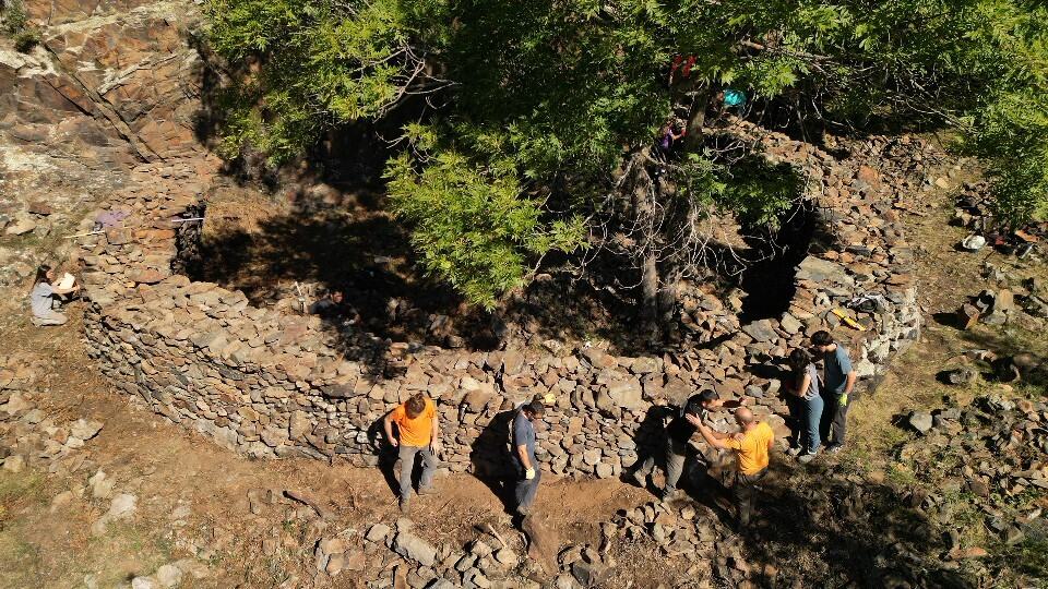 Un grupo de personas trabaja en torno a una estructura circular de piedra construida alrededor de un árbol en una zona rocosa al aire libre. Forma parte del patrimoni cultural arqueològic